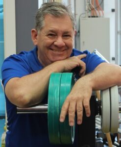 Smiling man in a blue polo leaning on weight plates in a gym setting.