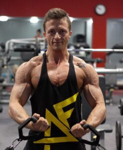 Muscular man in a black tank top with a yellow logo, gripping cable handles in a gym.