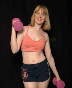 Woman in a coral sports bra and navy shorts lifts a pink dumbbell in a gym-like setting, smiling at the camera.