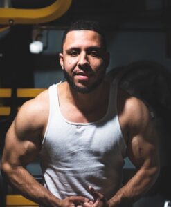 Athlete in a white sleeveless shirt flexing arms in a dimly lit gym with weights in the background