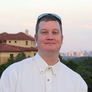 Man in a white button-down shirt with sunglasses on his head, outdoors with houses and a distant skyline behind him.