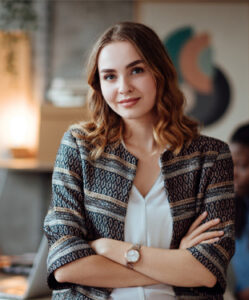 Confident young woman with wavy light brown hair, wearing a patterned jacket and white blouse, standing in an office with a blurred background.