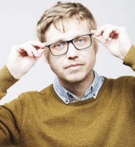 Man with light brown hair and stubble adjusts his black glasses, wearing a mustard sweater over a denim shirt.