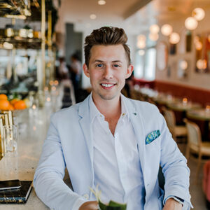 Smiling man in a light blue blazer sits at a bar with a drink in a bright, modern cafe setting behind him