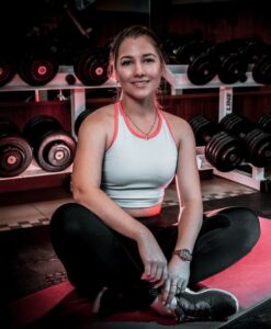 Athlete sits cross‑legged on a red gym mat, wearing a light blue tank top and black leggings, smiling with dumbbells on racks behind her.