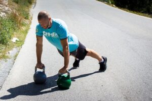 Man in a blue CrossFit shirt performing a push-up with two kettlebells on an outdoor road.