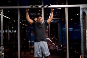 Man in a black shirt and gray shorts does a pull-up on a gym bar, arms extended above his head, in a dimly lit fitness center.