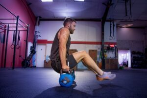 Man in a gym performing a kettlebell exercise on the floor, hands gripping a blue kettlebell with knees bent and torso leaning back.