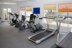Cardio gym with treadmills, stationary bikes, and an elliptical machine along windows with orange blinds in a bright room.