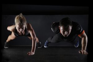 Two athletes performing push-ups on a dark gym floor, facing downward with arms extended.