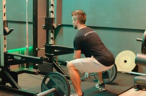 Man in a black shirt and gray shorts squats with a loaded barbell in a gym power rack on a lifting platform.