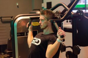Man in a gym uses a cable machine for a shoulder press exercise, gripping the handles.