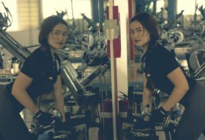 Woman in a gym wearing black workout clothes, bending with dumbbells in front of a full-length mirror by the weights area.
