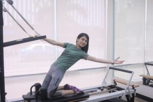 Woman wearing a teal shirt and gray leggings stretches sideways on a Pilates reformer in a bright gym room with large windows.