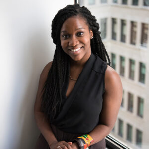 Professional portrait of a smiling Black woman with braided hair, wearing a black sleeveless top, standing by a window with a cityscape outside.