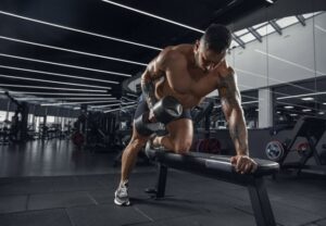 Muscular man shirtless doing a dumbbell row on a workout bench in a modern gym