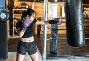 A woman wearing purple gloves throws a straight punch at a hanging punching bag in a gym.