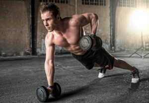 Muscular man in a gym performs a one-arm dumbbell row, propping on a dumbbell with a focused stance in a gritty, industrial setting.