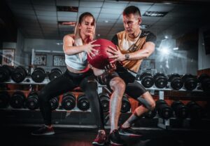Two teammates in a gym push a red medicine ball between them during a partner workout, with dumbbells lining the rack in the background.