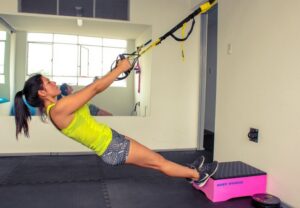 Woman in a neon-yellow top performs a suspended TRX row with feet on a pink box in a gym gym environment.