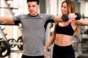 Man and woman in a gym performing dumbbell lateral raises with arms extended to the sides.
