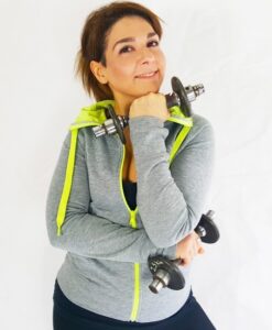 Woman in a gray zip-up hoodie with neon-yellow accents, smiling, holding dumbbells in a fitness pose.