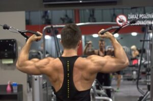 Bodybuilder in a gym viewed from behind, arms raised with cable handles for a back exercise.