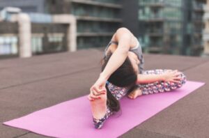 Person performing a yoga stretch on a pink mat outdoors on a rooftop, reaching toward their foot with buildings in the background on a cloudy day.