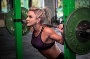 Woman in a purple sports bra squats with a loaded barbell inside a green squat rack at the gym, muscles tensed and focused on the lift.