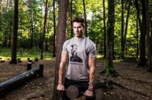 Man wearing a gray T-shirt lifts a log in a sunlit forest, surrounded by trees and fallen leaves, mid-workout.