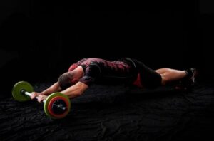 Male athlete in a black-and-red outfit lies face down on a dark mat, performing a prone barbell row with a barbell loaded with bright green weight plates.