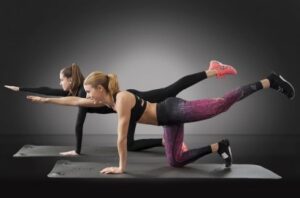 Two women in athletic wear perform a stability exercise on exercise mats in a studio; they balance with opposite arm and leg extended.