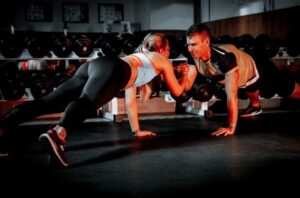 Two athletes in a gym perform a partner push-up facing each other, with dumbbells and equipment in the background of the weight room.