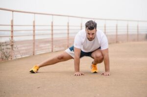 Man in a white shirt and orange shoes performs a stretch on a boardwalk with a railing by the water.