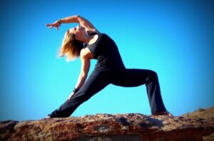Woman practicing yoga in a warrior-like pose on a rocky ledge against a bright blue sky.