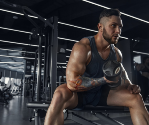 Muscular man in a blue sleeveless shirt curls a dumbbell on a gym bench.