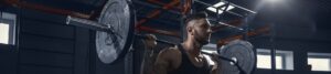 Man with a tattooed arm in a black tank top lifting a heavy barbell on a squat rack in a gym (back squat).