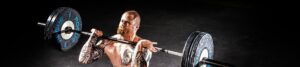 Muscular tattooed man lifting a heavy barbell in a dark gym.