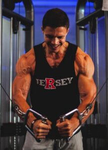 Muscular man in a sleeveless black shirt gripping cable handles in a gym, smiling as he performs a workout.