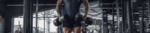 Person in a gym standing with two heavy dumbbells at their sides, back to camera, wearing a dark shirt and black shorts.