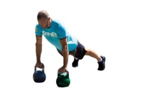 Man in a turquoise T-shirt performing a two-kettlebell exercise in a partially extended plank position, with one leg raised behind him, on a white background.