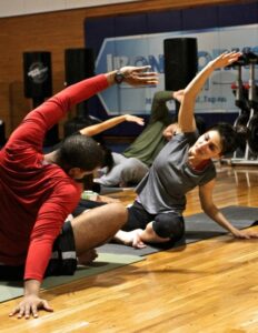 People in a gym doing partner stretches on yoga mats, with a man in red reaching his arm overhead and a woman in gray following the pose.
