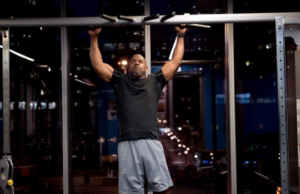 Man in a black shirt and gray shorts performs a pull-up on a gym bar at night, arms raised overhead.