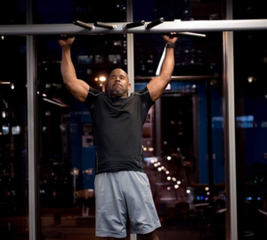 Man in a black t-shirt and gray shorts performs a pull-up on a gym bar with city lights visible through large windows behind him.
