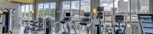 Bright gym with treadmills and cardio machines facing large windows overlooking city skyline outside the building's glass wall(s).