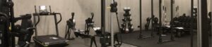 Industrial gym interior with treadmills, stationary bikes, weight benches, and a rack of dumbbells and barbells along a concrete wall.