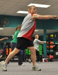 Man in a red and gray sleeveless shirt punches forward in a group fitness class at the gym, with other participants and equipment in the background while he lunges.