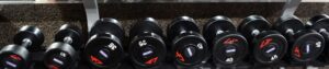 Row of black dumbbells on a weight rack in a gym, with speckled flooring in the background.