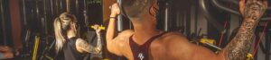 Person with diamond tattoos on arms performs a lat pulldown in a gym while another person with a ponytail observes nearby, both focused on workout equipment