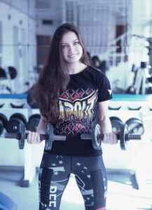 Young woman in a gym standing and lifting two dumbbells, smiling at the camera.